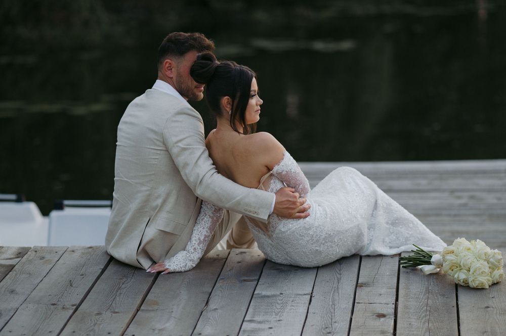 Newlyweds share an intimate moment sitting together on a wooden dock at their wedding.