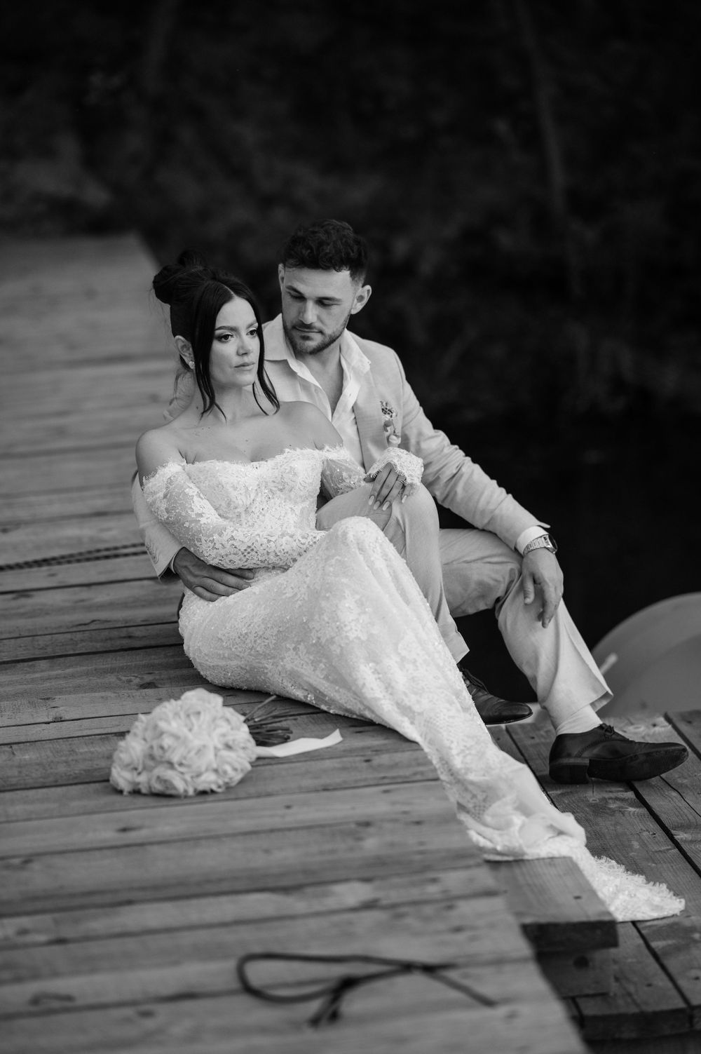 A romantic black and white wedding photo of a couple sitting intimately on a wooden dock.