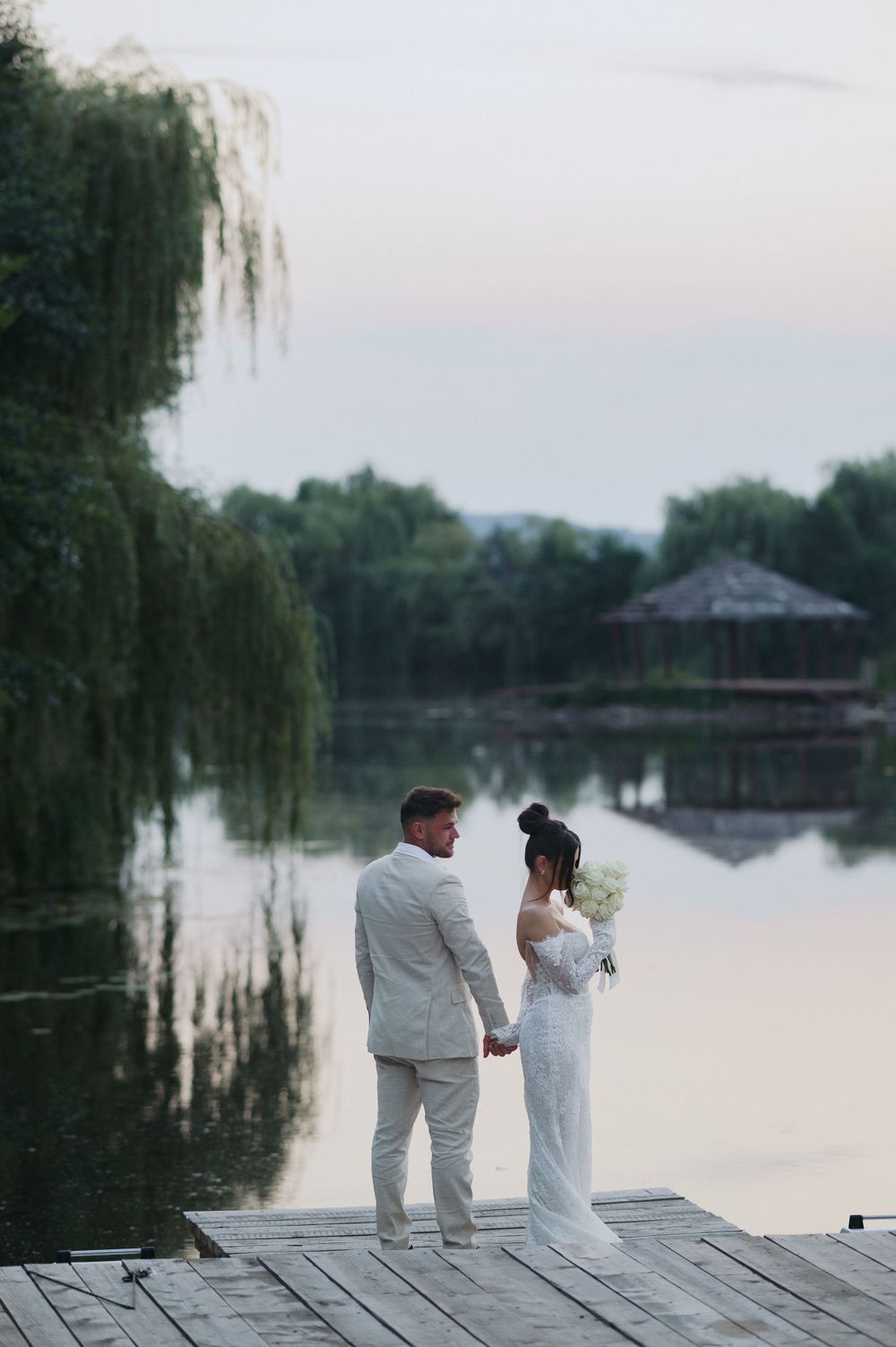 A couple stands on a wooden dock overlooking a tranquil lake at sunset with weeping willow trees in the background.
