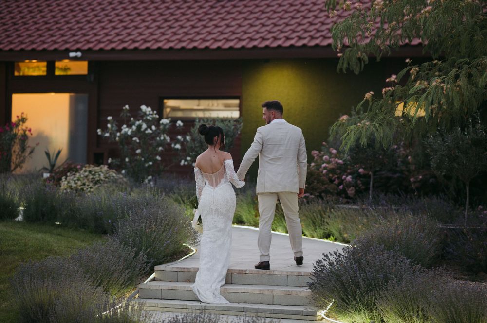 A couple in white attire stand together on garden steps at dusk with lavender plants and warm lighting in background.