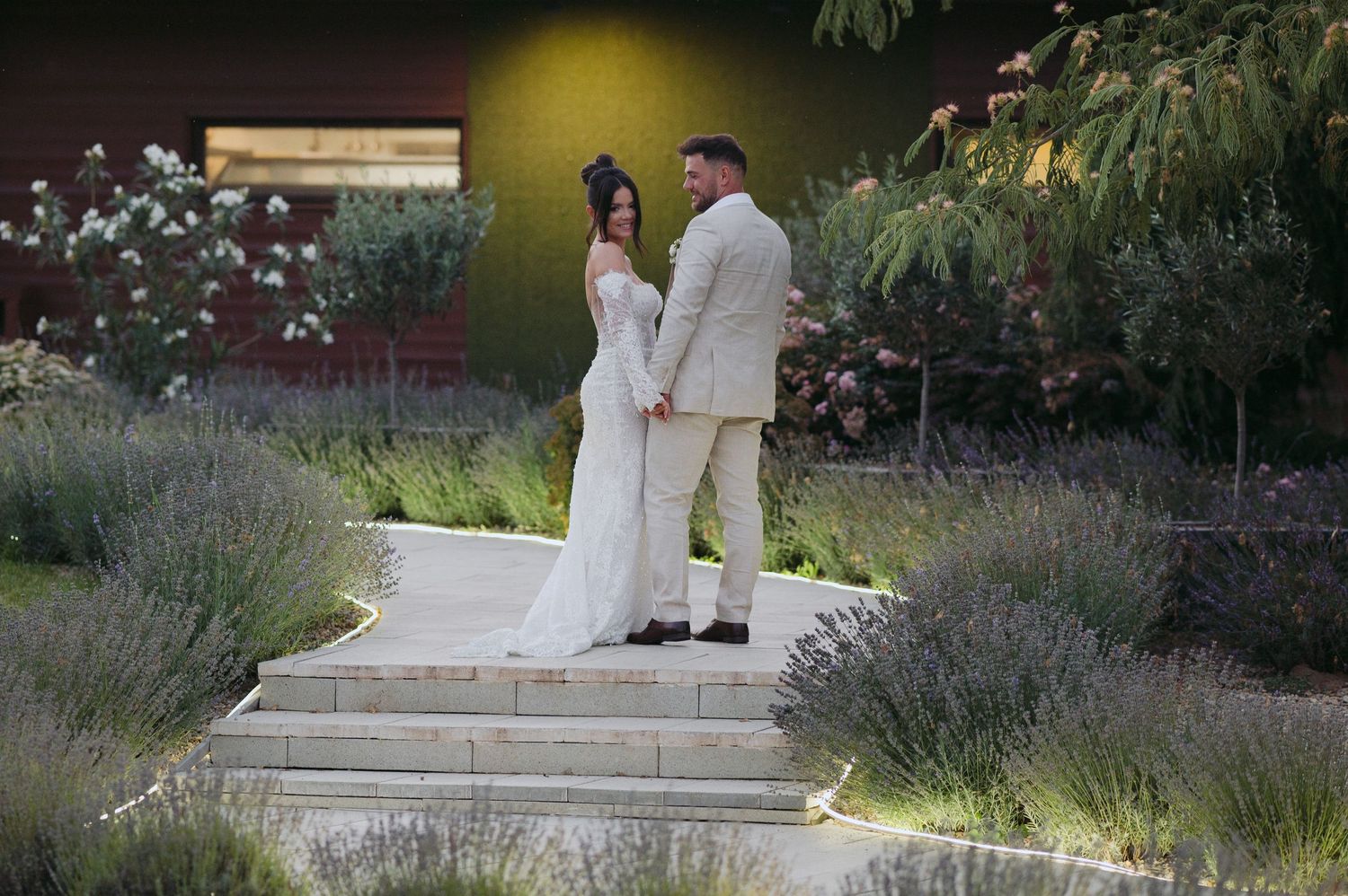 Couple in wedding attire stand on stone path surrounded by lavender plants and garden landscaping at sunset.