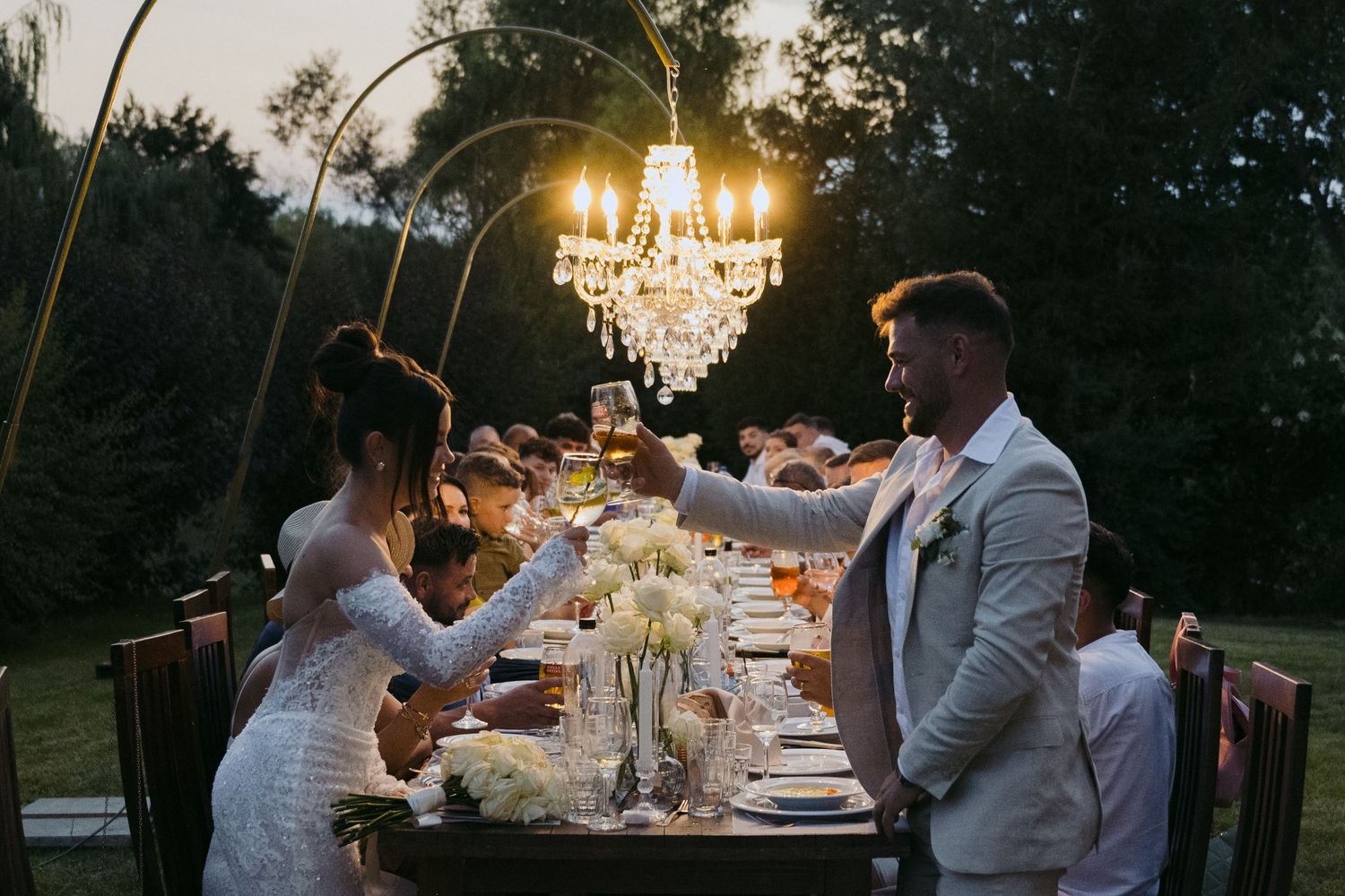 Elegant outdoor wedding reception table set beneath illuminated crystal chandelier at sunset with floral centerpieces.