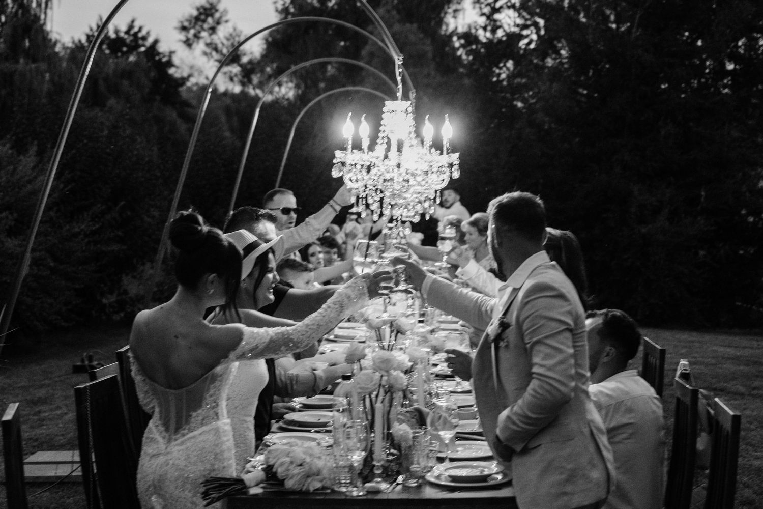 Elegant outdoor dinner party under a crystal chandelier with guests toasting at a long table in black and white.