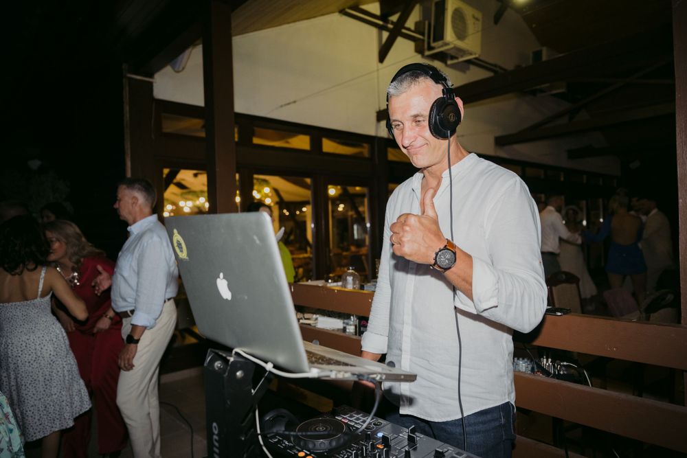 DJ performs at a nighttime event using a MacBook and wearing white headphones while standing at wooden tables.