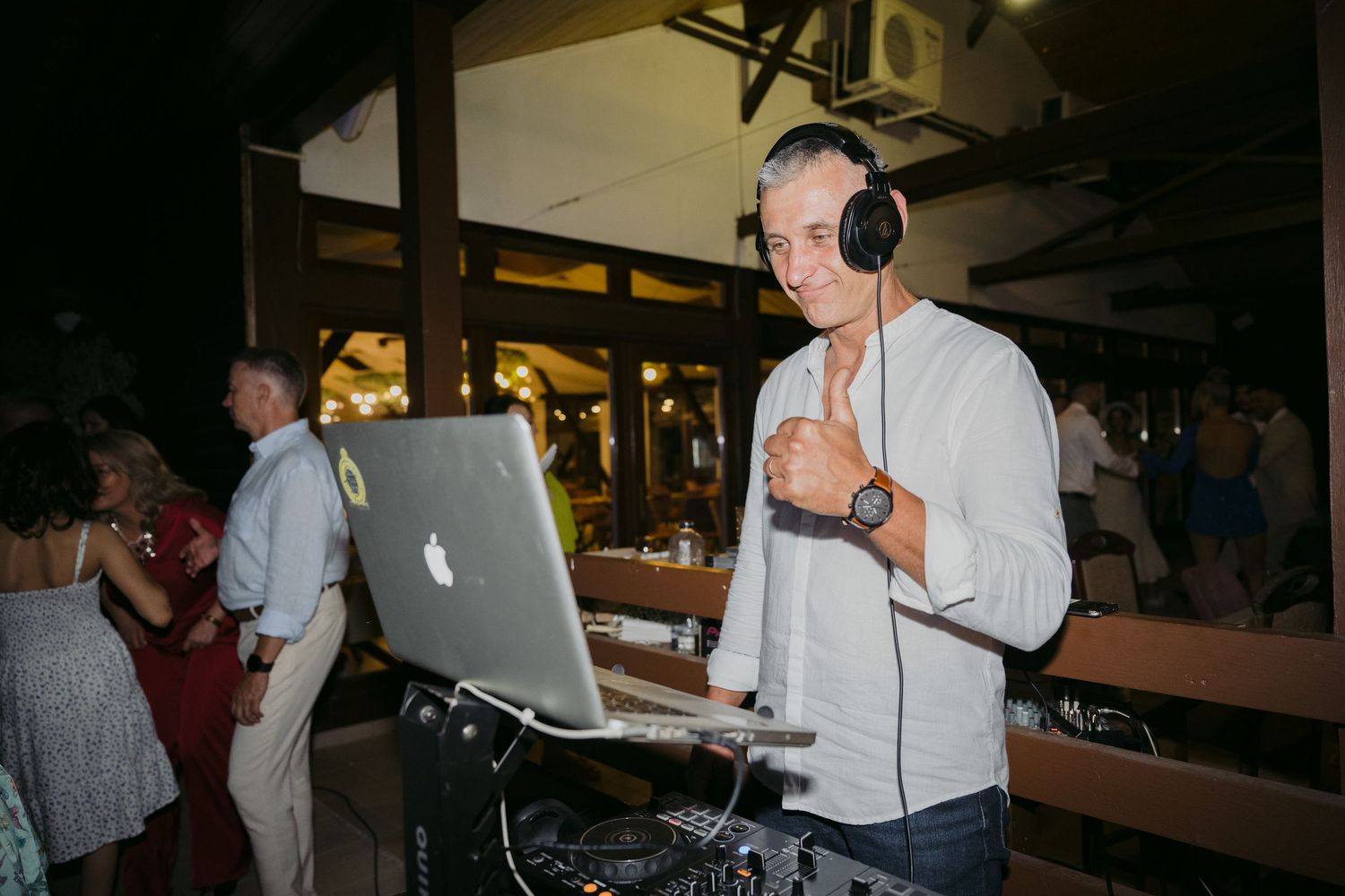 DJ performs at a nighttime event using a MacBook and wearing white headphones while standing at wooden tables.