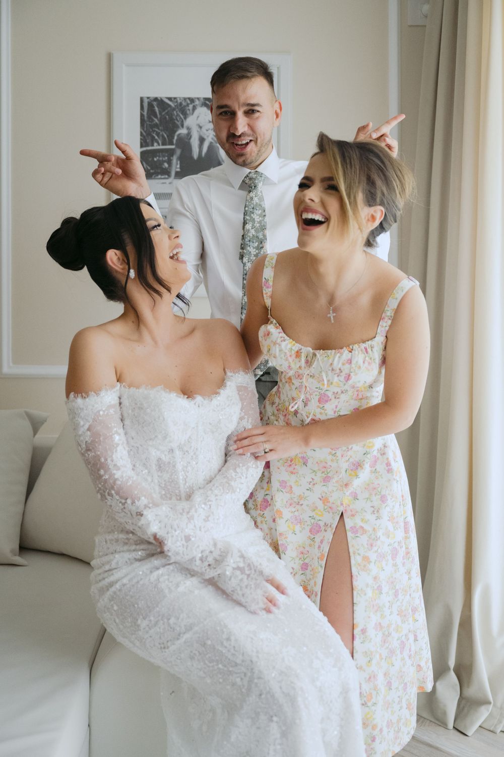 A bride in a white lace gown shares a joyful moment with wedding party members in a light-filled room.
