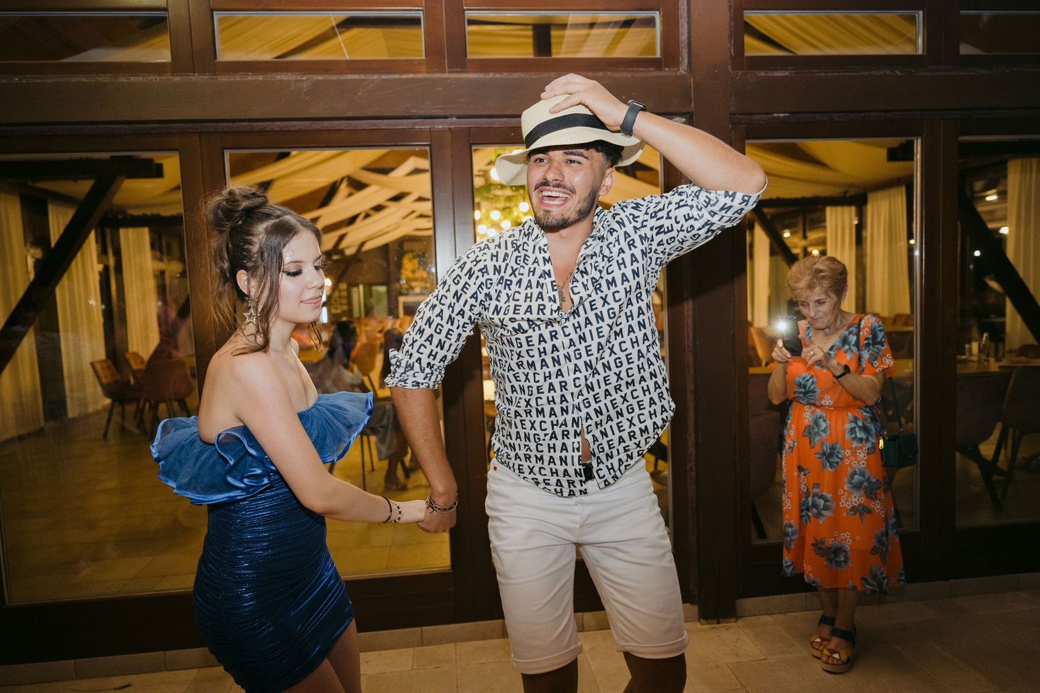 A couple dances energetically at a rustic indoor venue, with the person in a patterned shirt and hat moving animatedly.