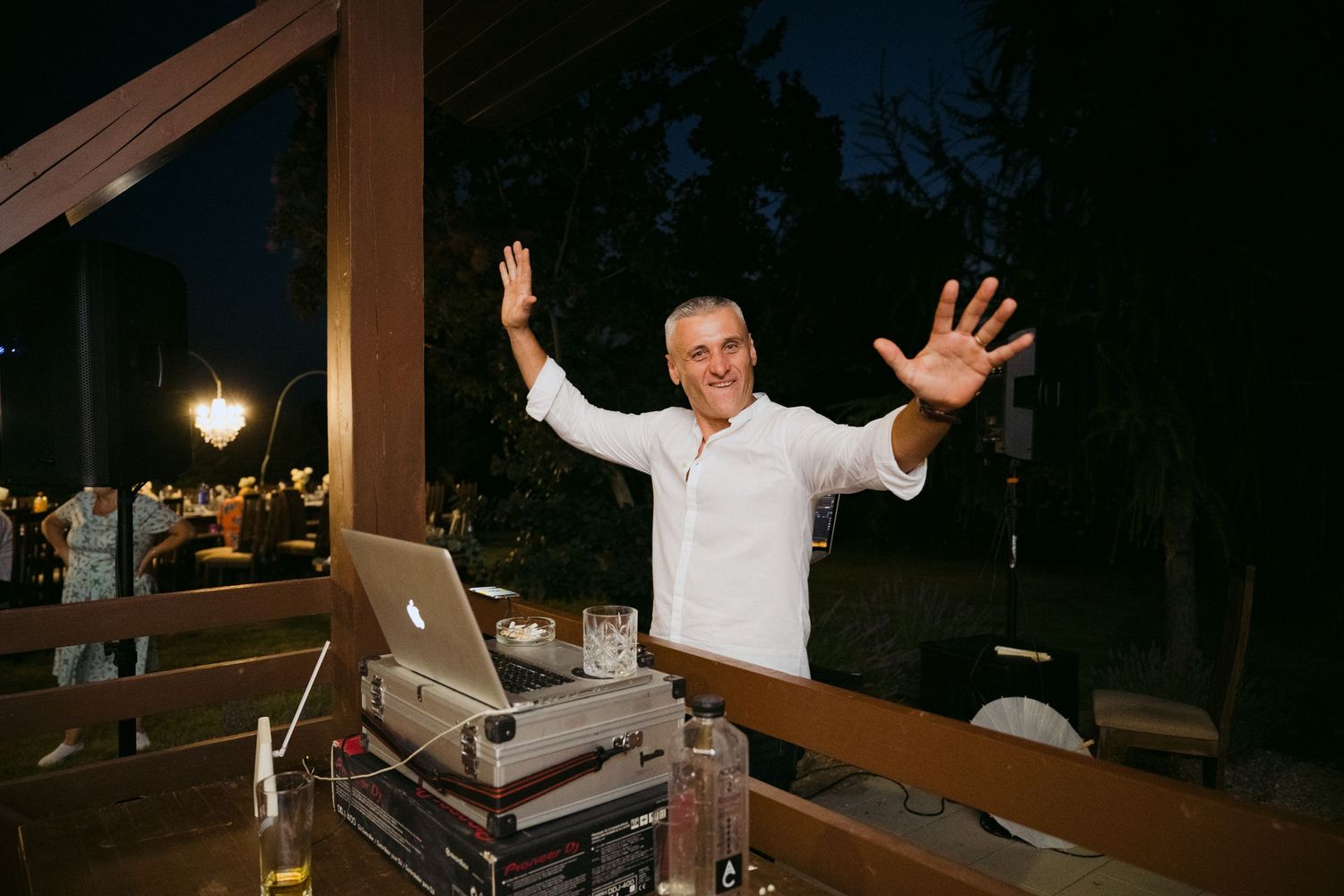 DJ stands with raised arms behind equipment setup at outdoor nighttime venue.