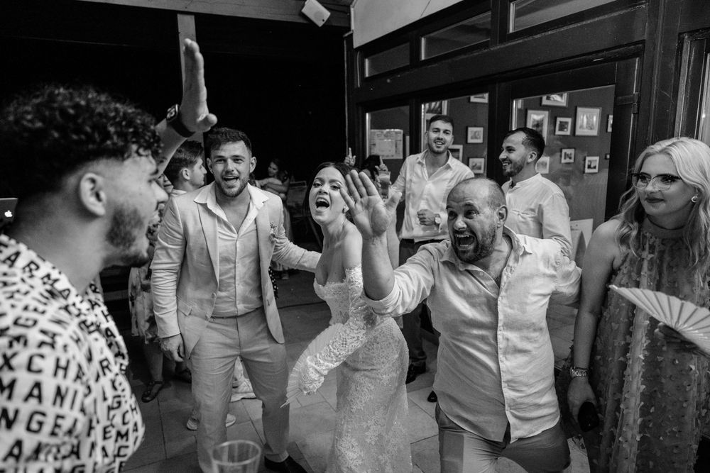 Black and white photo of a group of guests laughing and dancing together at a joyful wedding celebration.
