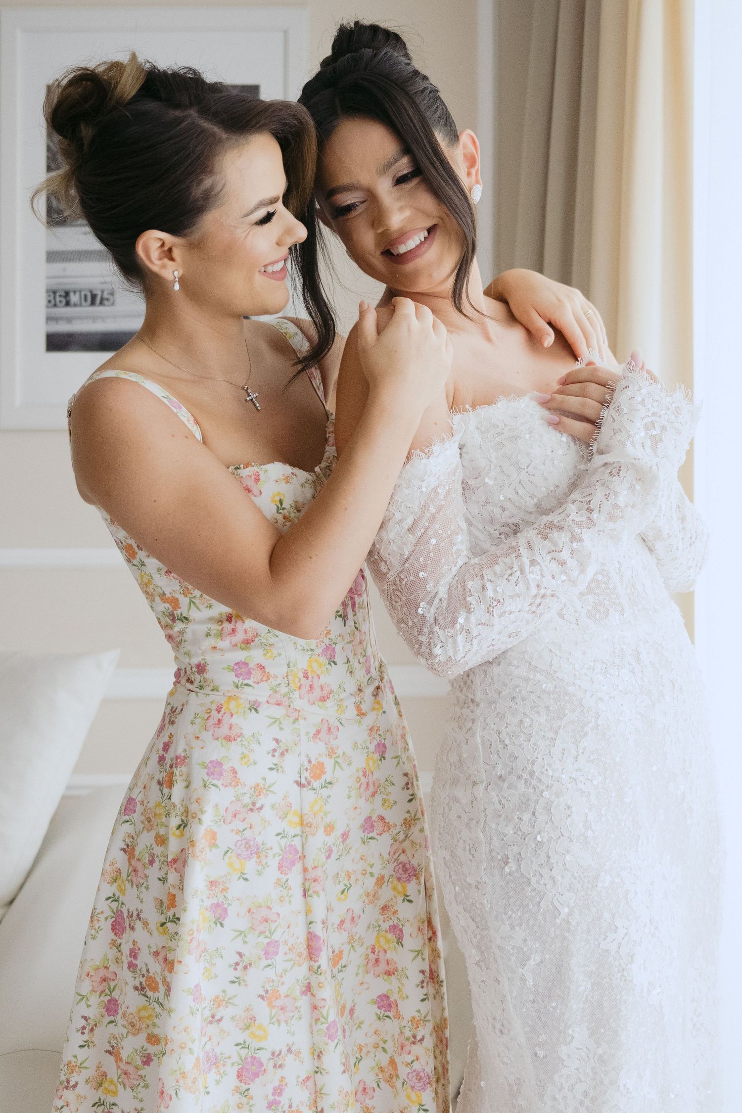 Friends share a joyful moment as one in a floral dress helps adjust the lace details on a white wedding gown.
