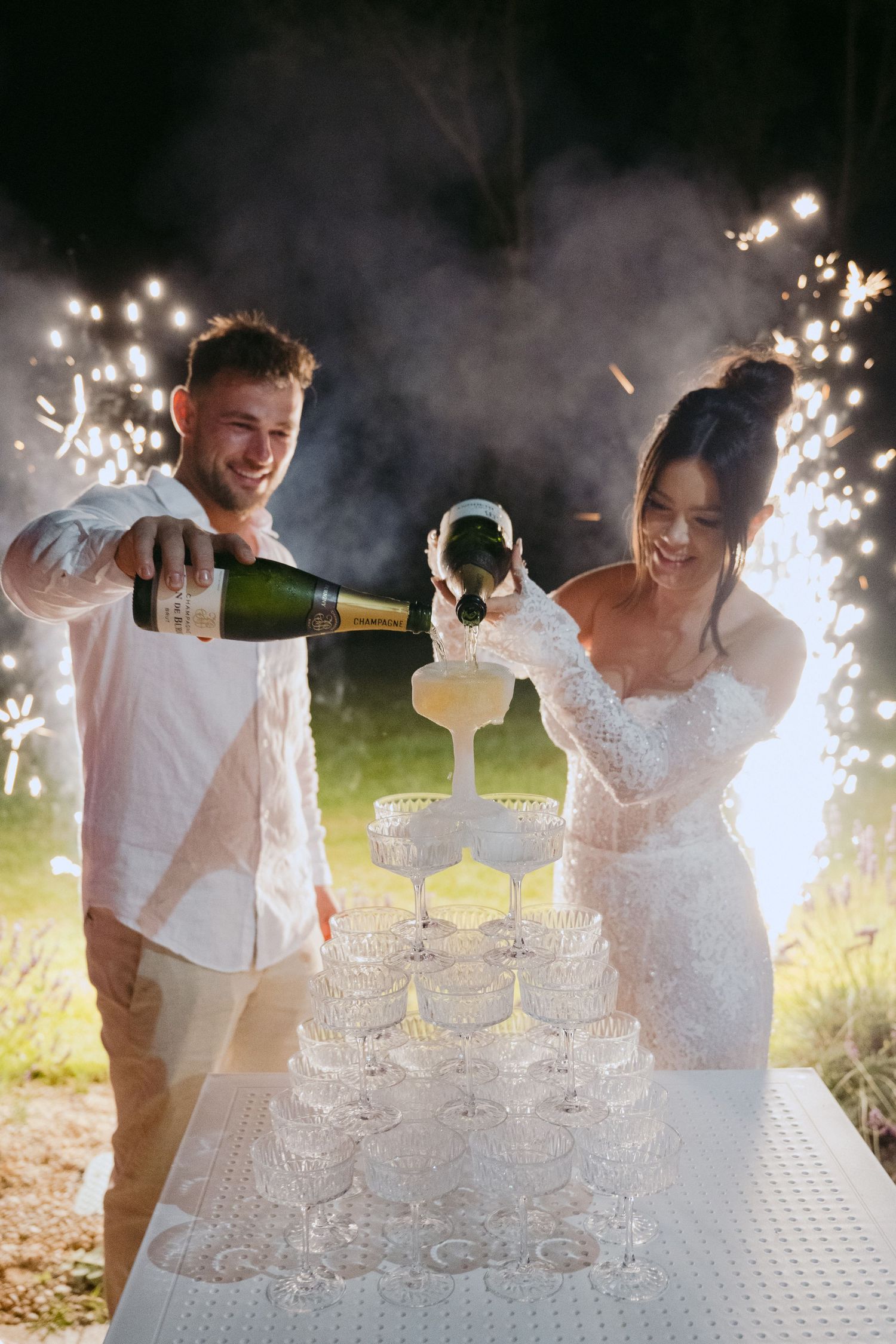 Couple pouring champagne into tower of glasses with sparklers creating magical wedding moment.