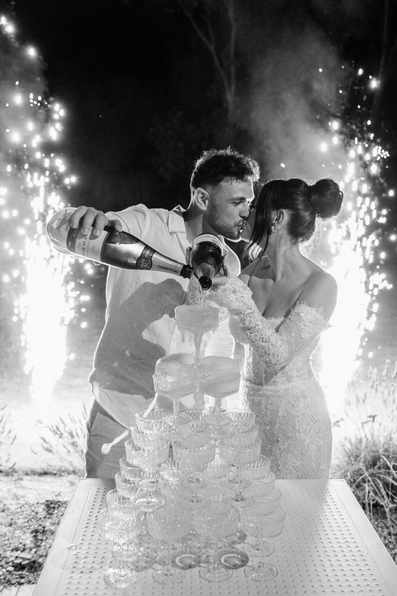 Newlyweds pour champagne into a tower of glasses with sparklers glowing behind them in dramatic black and white.