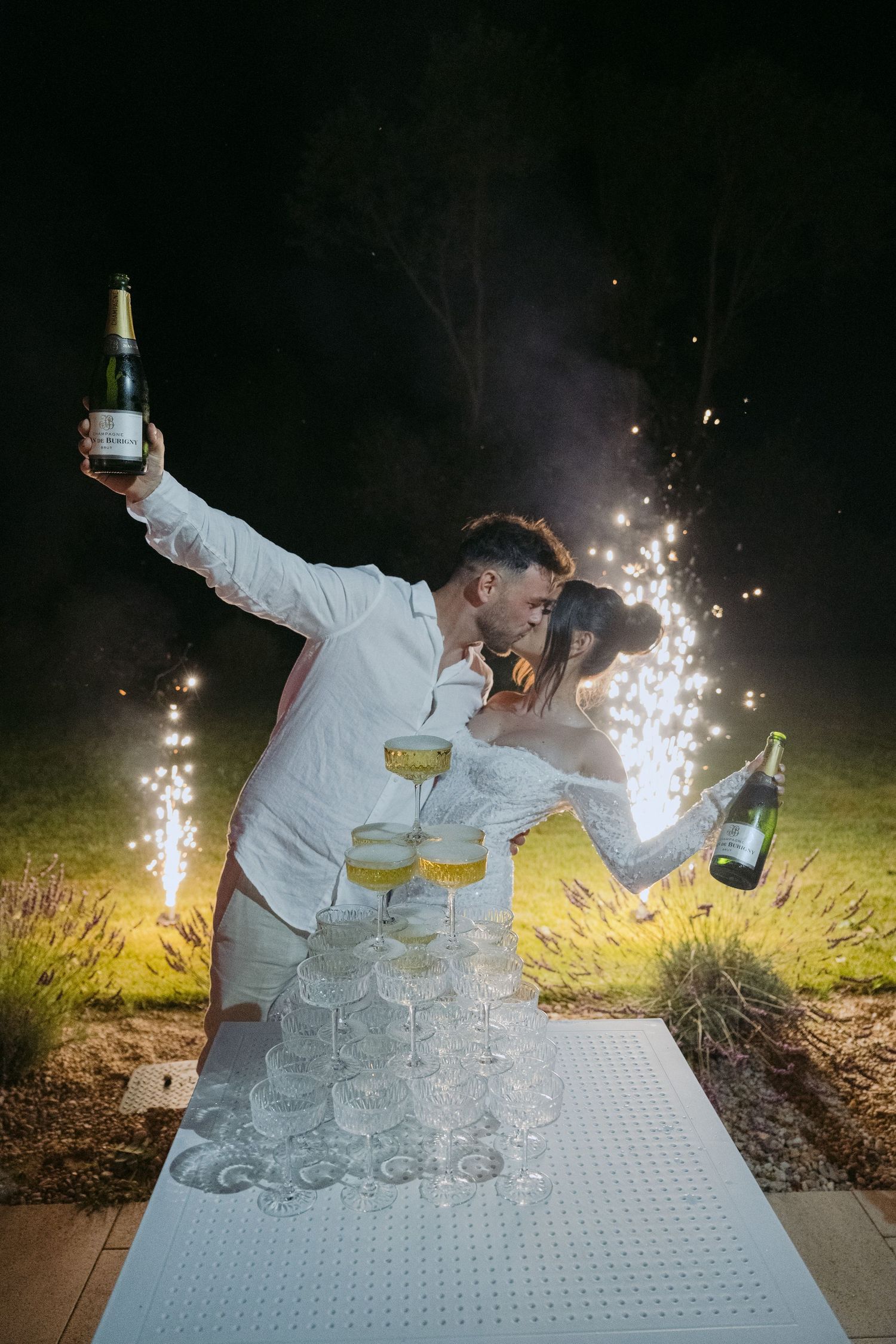 A celebratory moment with champagne tower and sparklers glowing behind a couple pouring at an outdoor evening event.