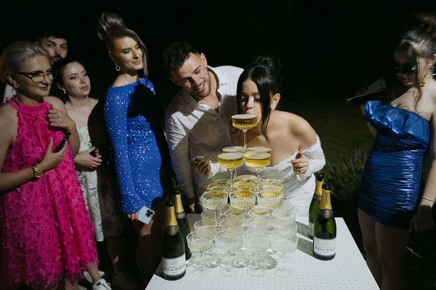 Party guests in colorful formal dresses gather around a table with champagne glasses and bottles during a celebration.