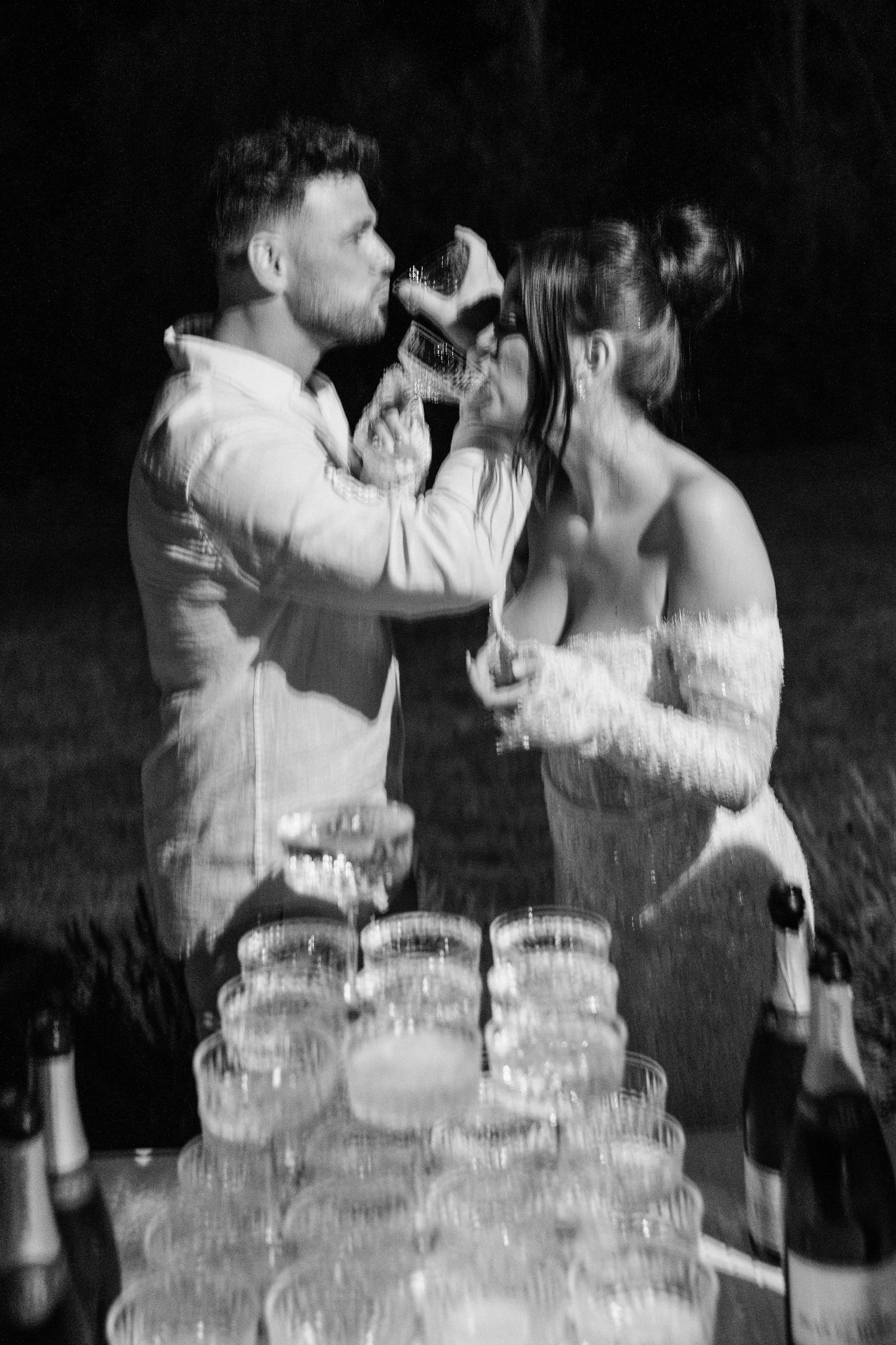 A couple in wedding attire shares a champagne toast next to a tower of stacked glasses at their reception.
