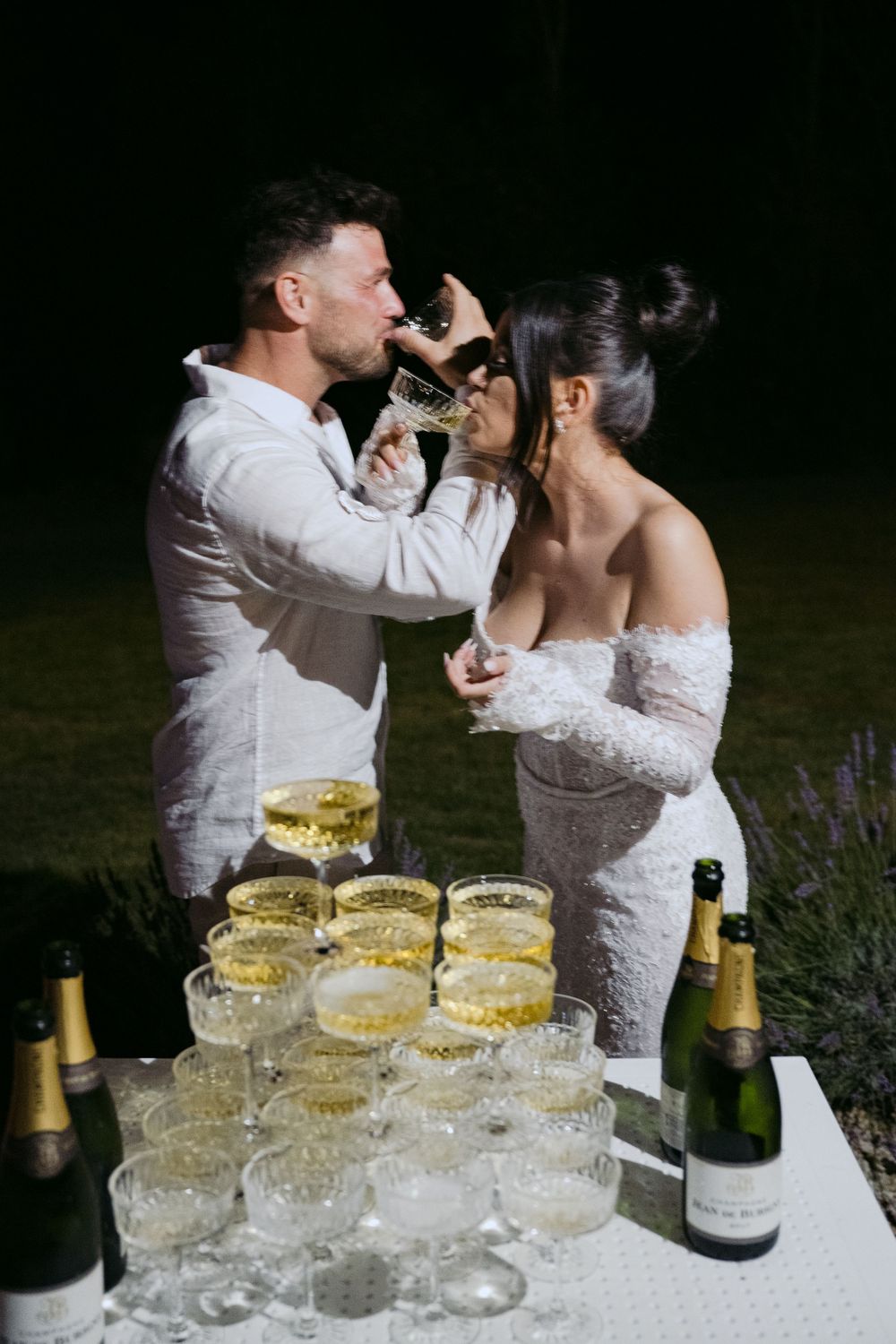 A couple in white wedding attire share champagne together at a table filled with elegant champagne glasses.