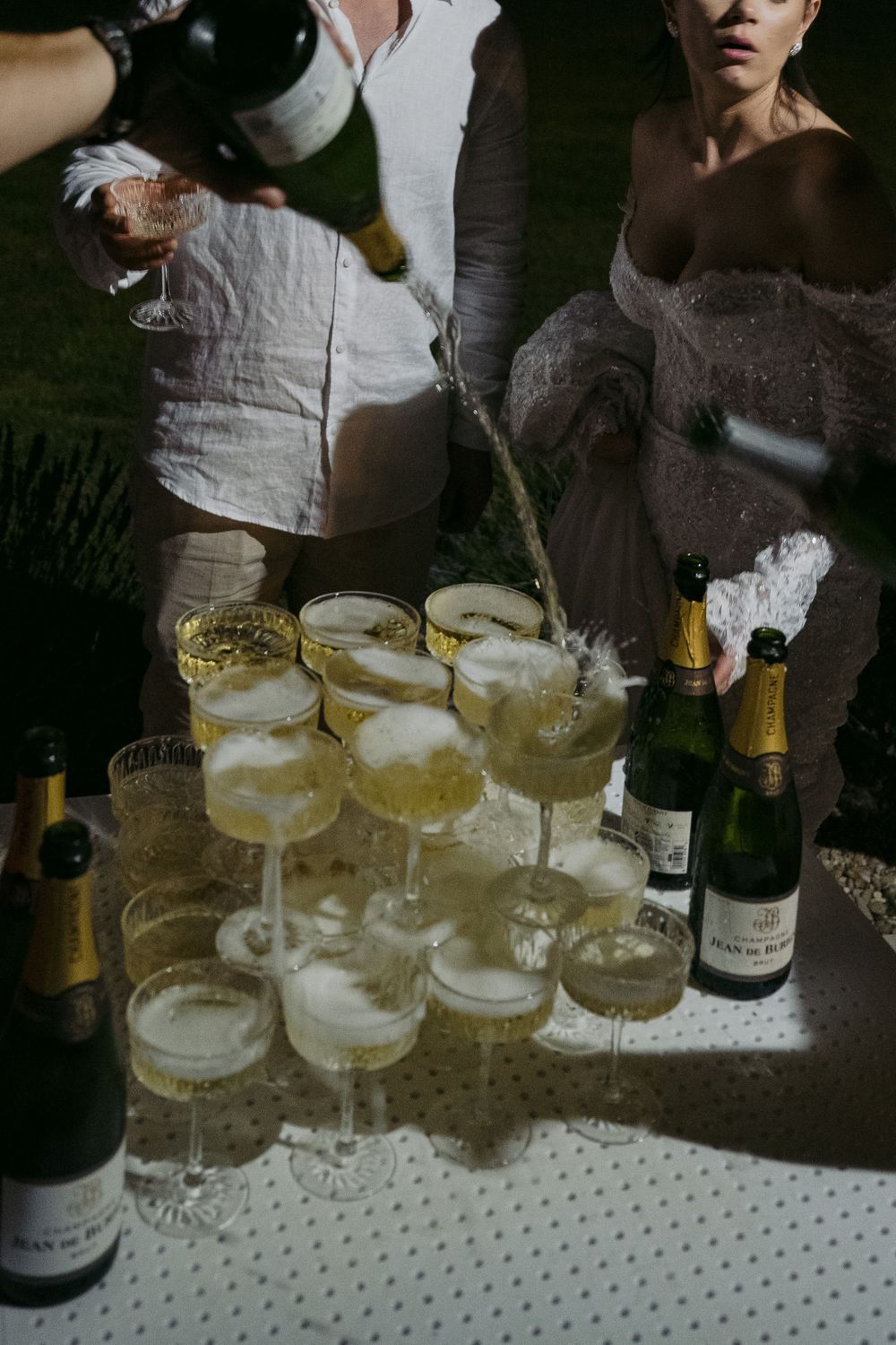 Champagne being poured into a tower of elegant coupe glasses on a white tablecloth during a celebration.