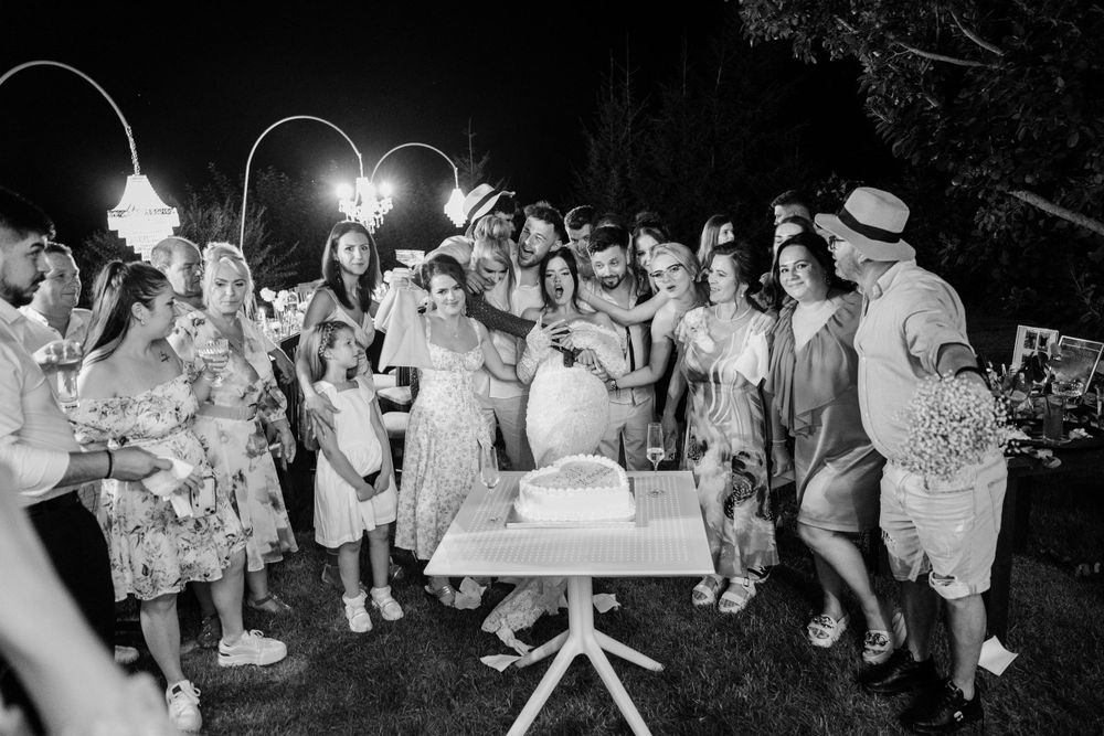 An outdoor evening celebration with a group gathered around a table under string lights in black and white.