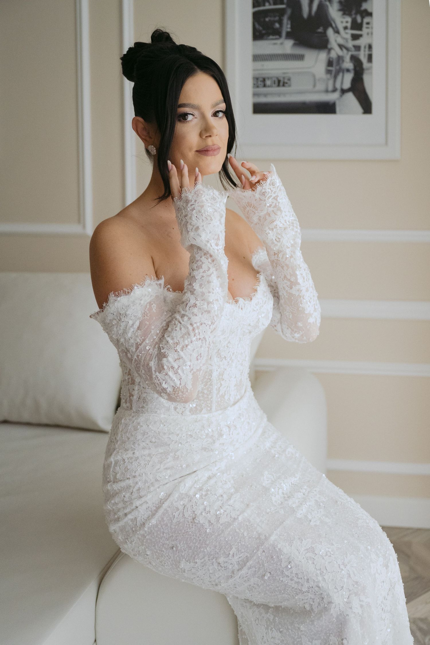 Bride wearing fitted white lace wedding gown with off-shoulder design and sheer long sleeves poses on white couch.