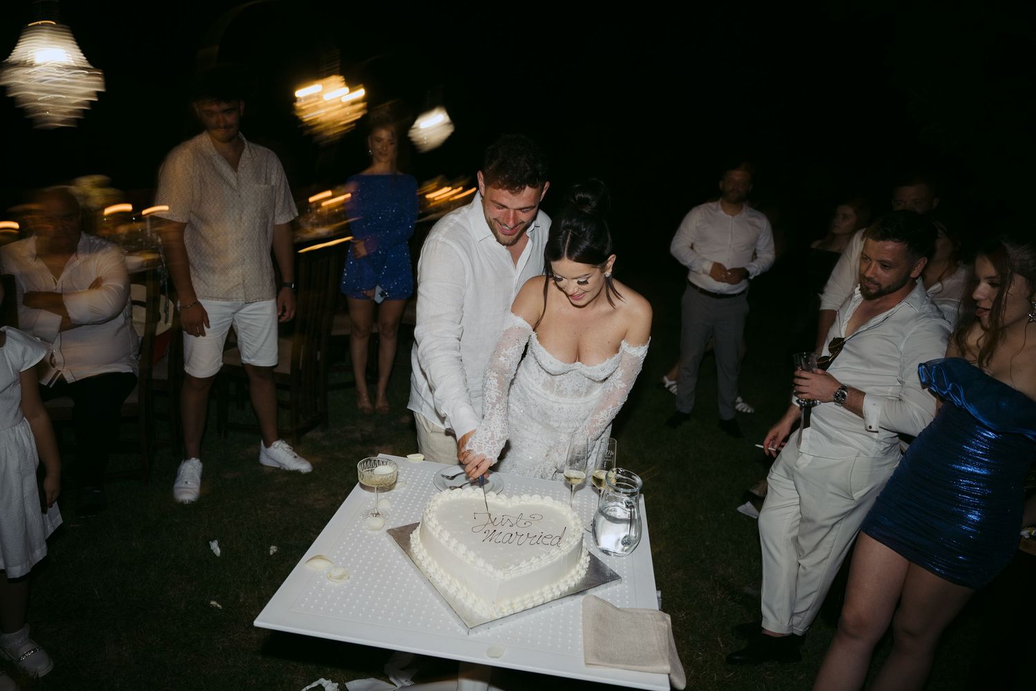A wedding couple cuts their heart-shaped cake at an outdoor nighttime reception.