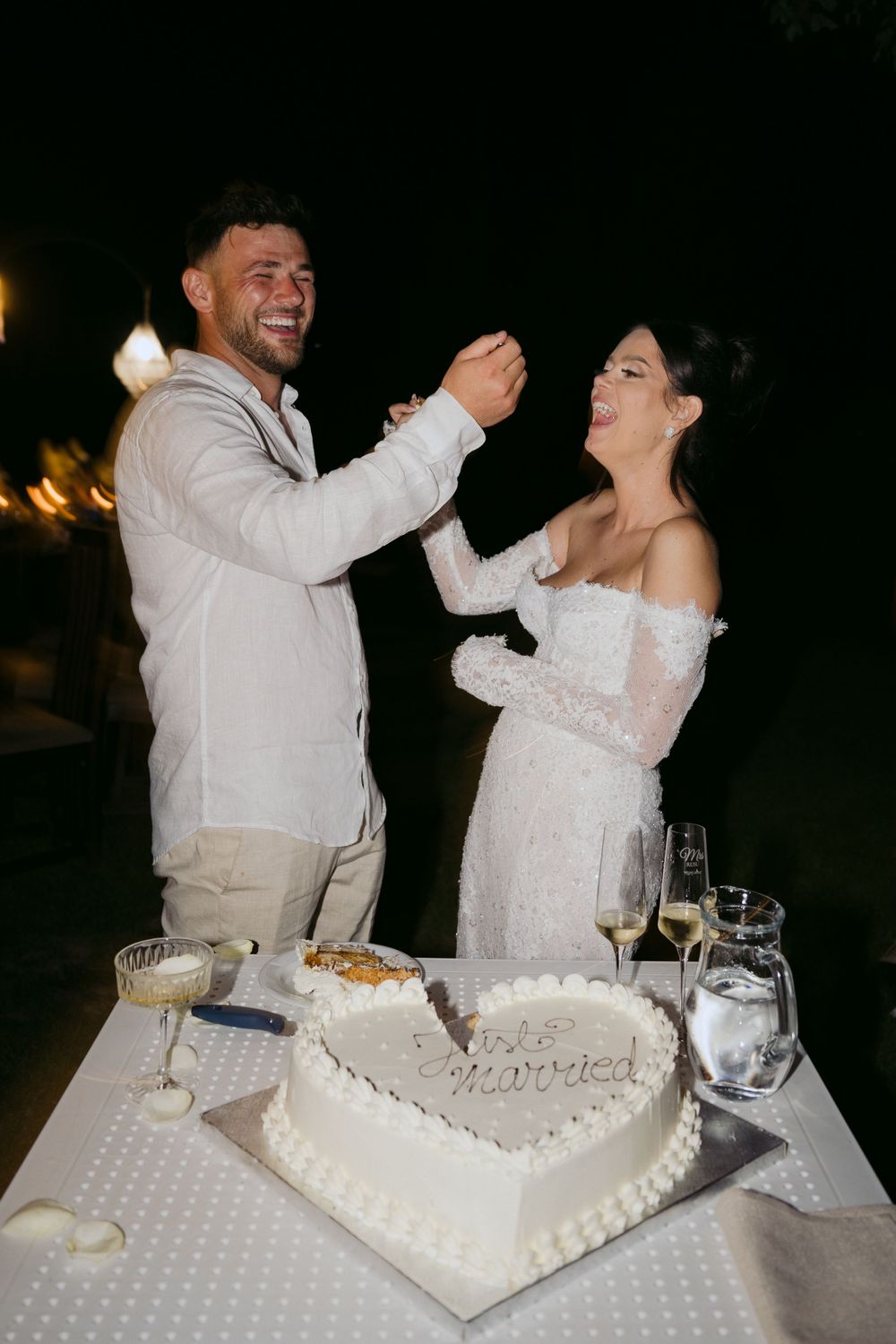 Newlywed couple sharing traditional cake feeding moment during outdoor evening reception with heart-shaped cake on table.