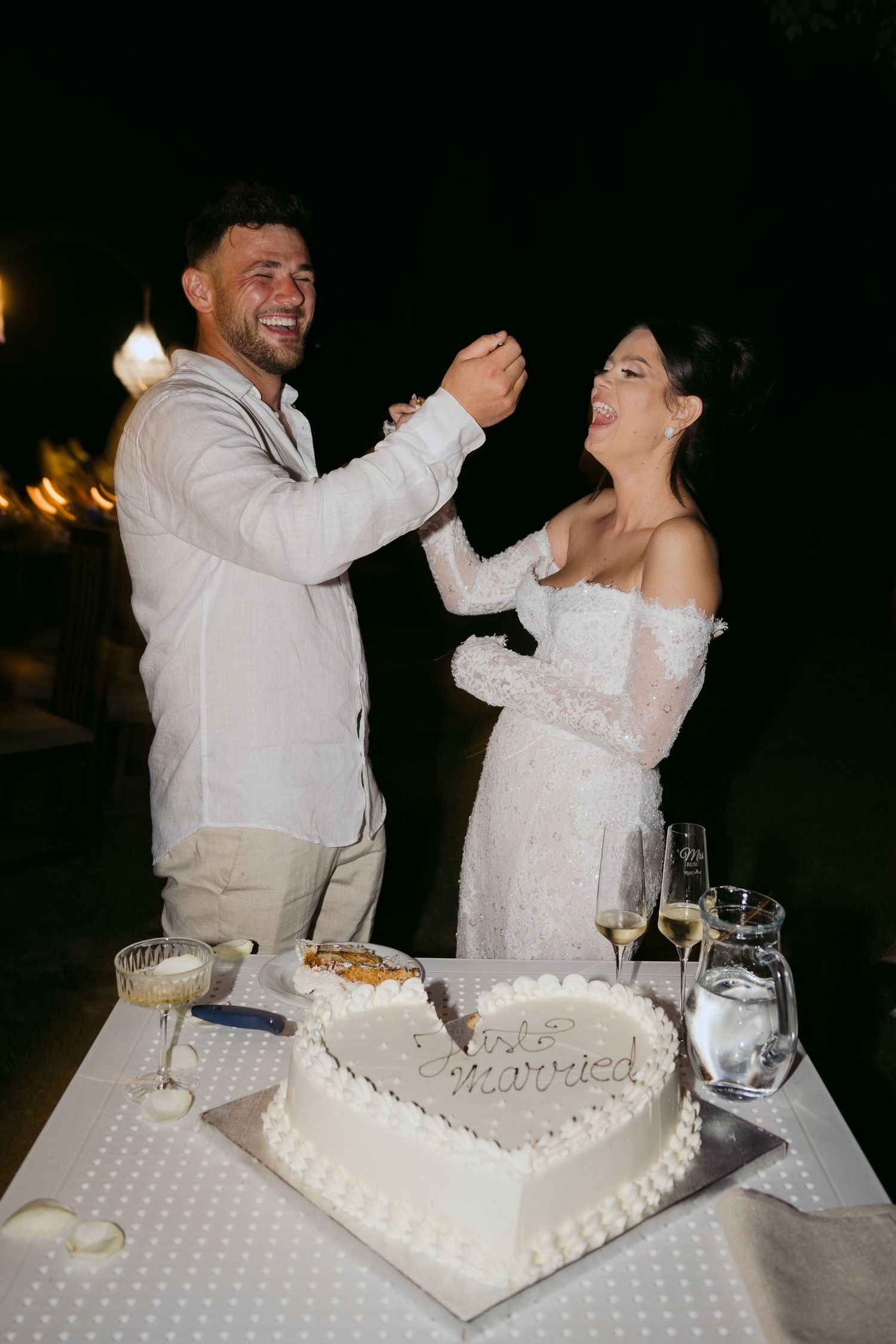 Newlywed couple sharing traditional cake feeding moment during outdoor evening reception with heart-shaped cake on table.