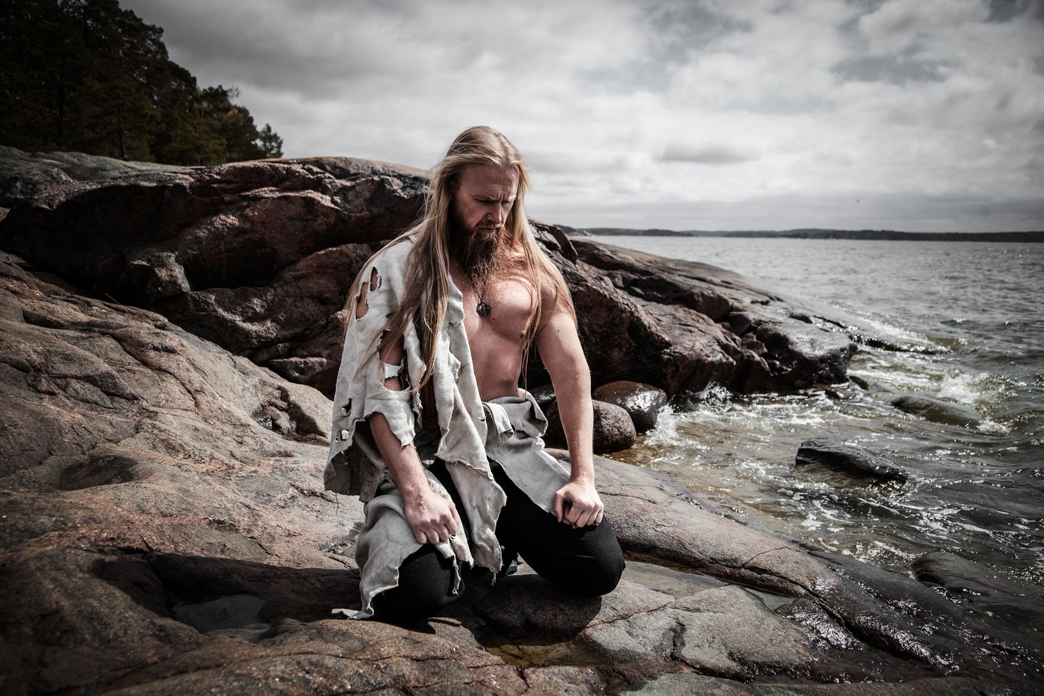 Person with long hair sits on rocky coastal shoreline on cloudy dramatic day by the ocean.