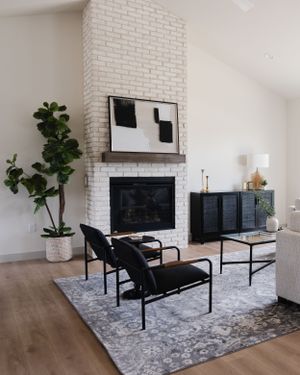 Modern living room with white brick fireplace, black furniture, abstract art, and a fiddle leaf fig plant on hardwood floors.
