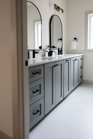 Modern grey bathroom vanity with double sinks, arched mirrors, black hardware and white countertop in a bright space.
