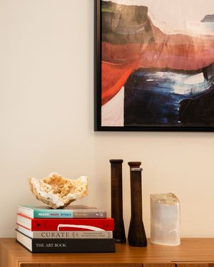 Modern home decor display with abstract wall art, stacked art books, dark candlesticks and a glass jar on wooden surface.