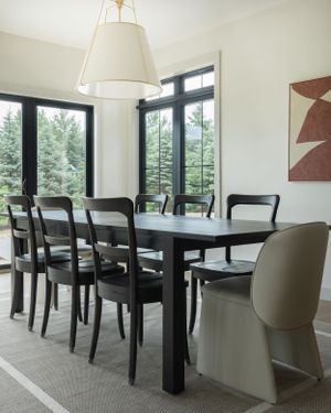 Modern dining room features black wooden chairs around dark table with large windows overlooking evergreen forest view.