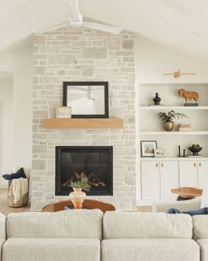 Modern farmhouse living room with whitewashed stone fireplace, beige couch, and built-in shelving displaying decorative accessories.