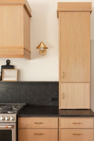 Modern kitchen with light wood cabinets, black countertops, and brass wall sconce against white walls.