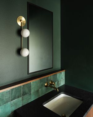 Dark green bathroom with brass fixtures, globe sconce, and emerald tile backsplash above undermount sink.