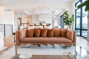 Modern living room interior with a tan leather sofa, geometric chandelier, and tall fiddle leaf fig plant by window.