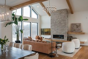 Modern living room features tall windows, stone fireplace, vaulted ceiling, and contemporary furnishings in neutral tones.