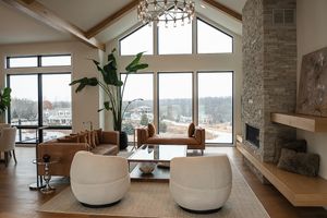 Modern living room with vaulted ceiling, stone fireplace, white poufs and panoramic windows overlooking snowy landscape.
