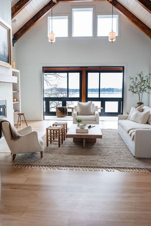 Modern coastal living room with vaulted ceiling, white furniture, and large windows overlooking waterfront view.