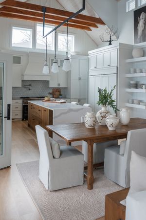 Bright open kitchen and dining area with exposed beams, white cabinets, and wooden dining table.