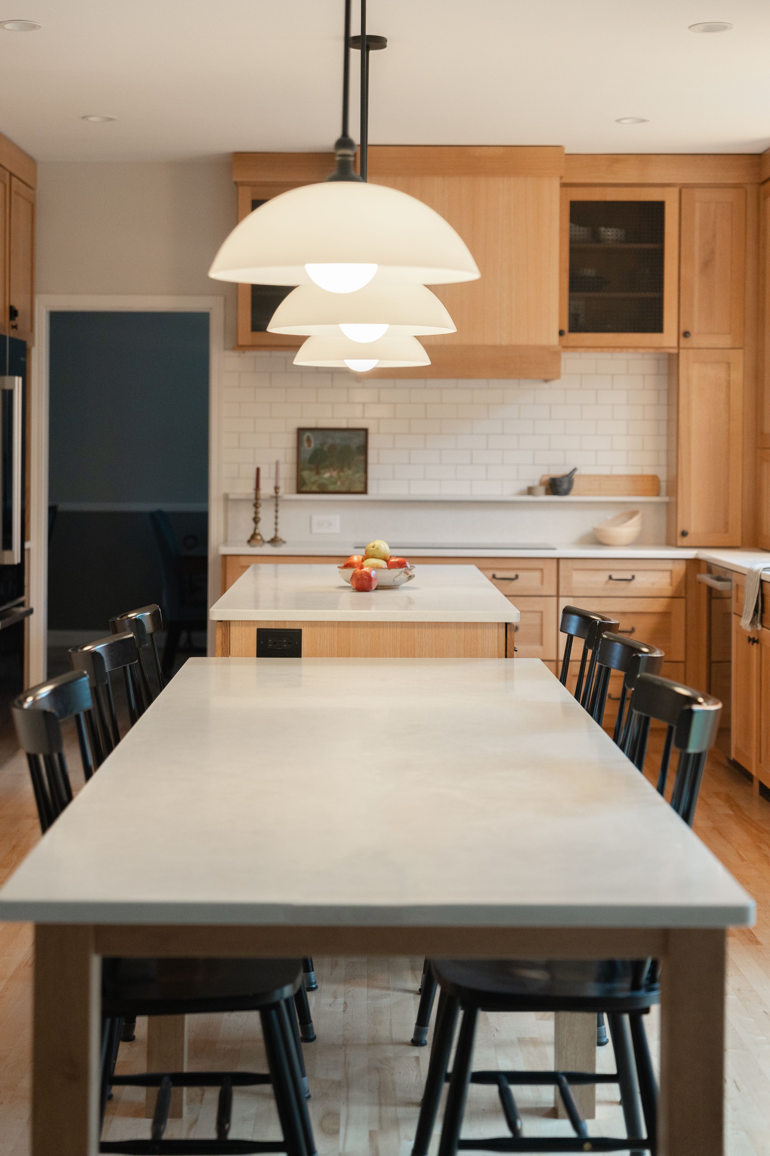 Modern kitchen featuring light wood cabinets, white subway tile backsplash, and a dining area with black chairs under a tiered pendant lamp.