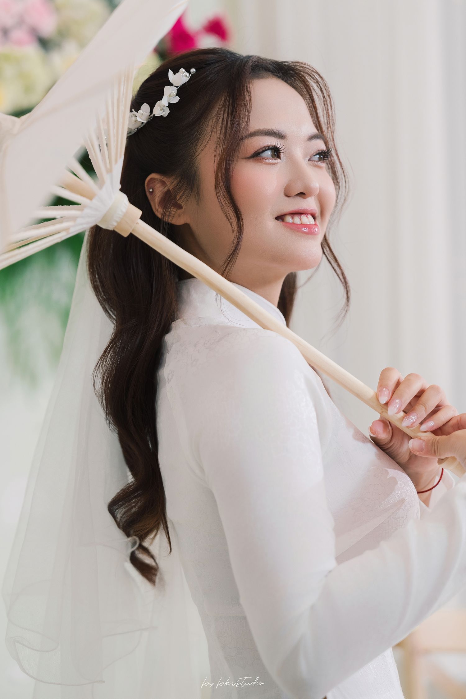 Traditional Vietnamese bride in white ao dai holding paper fan poses with gentle smile and delicate floral headpiece.