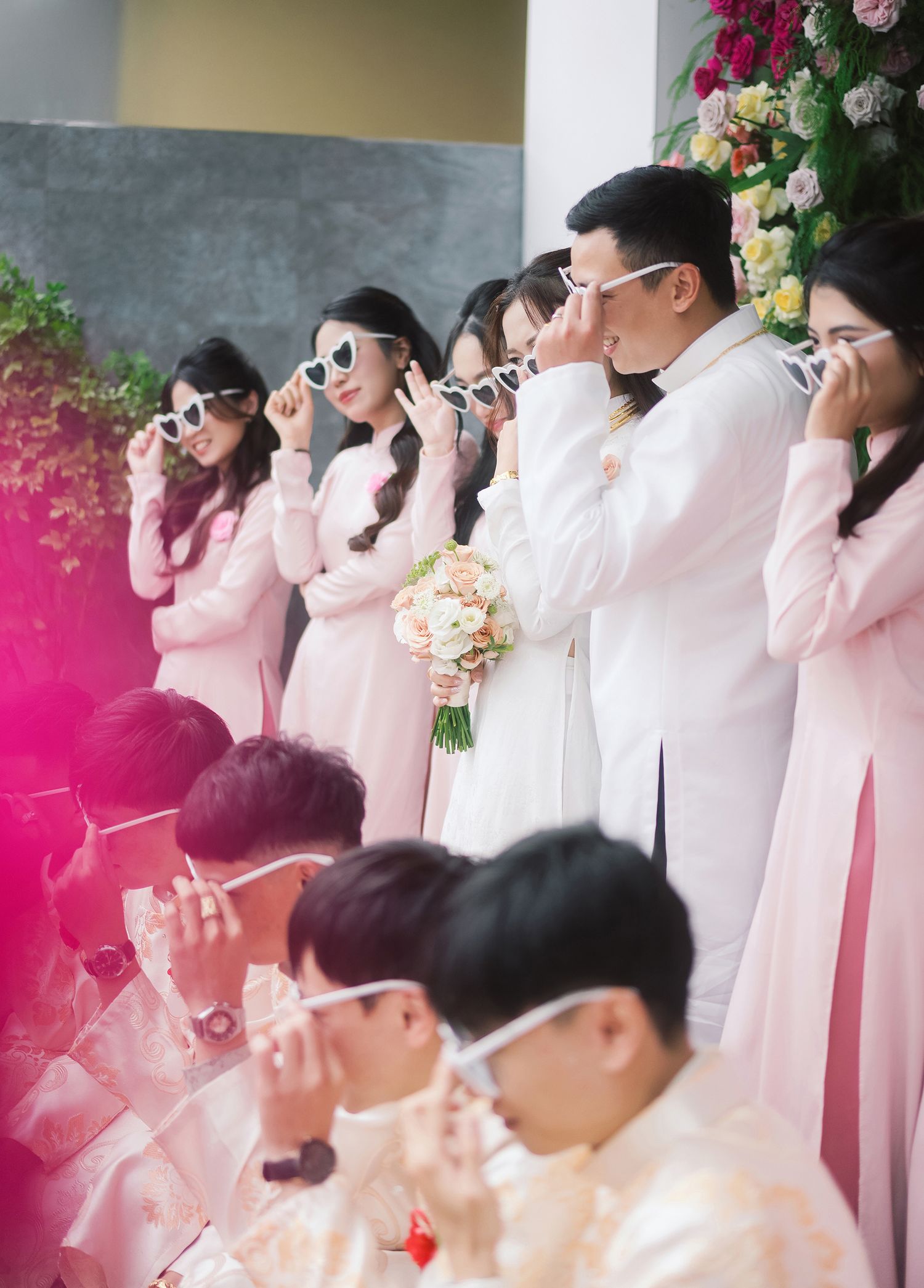 Wedding party group in matching pink outfits and sunglasses pose together for a photo surrounded by floral decorations.
