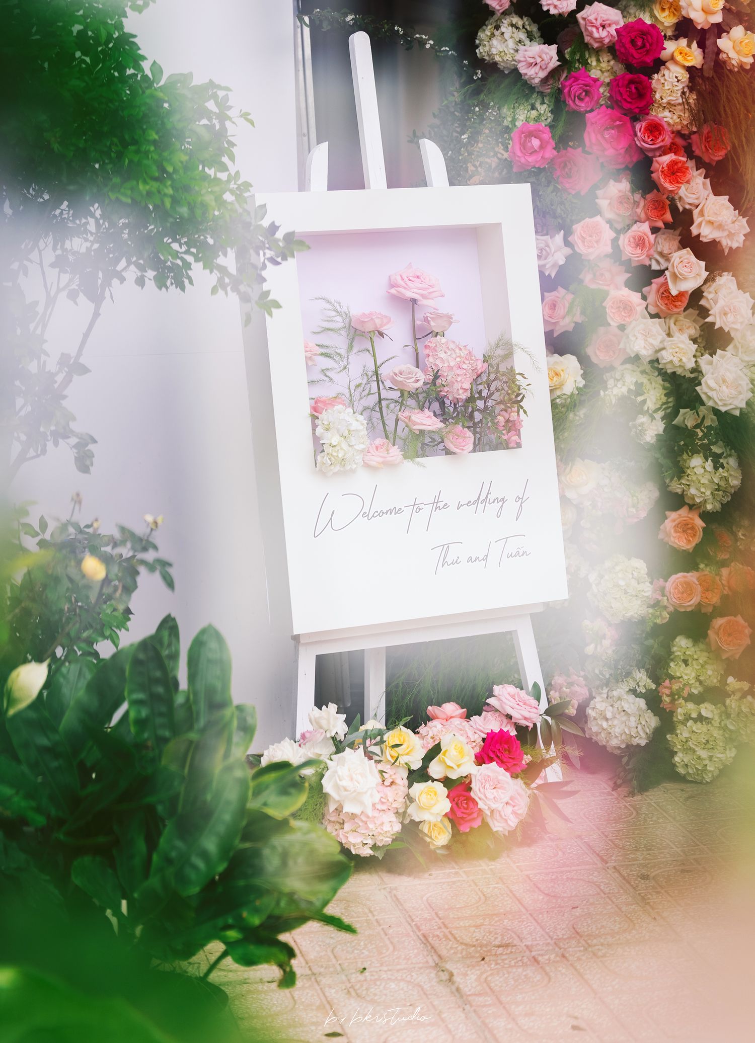 A white easel displays a floral photograph surrounded by pink and white roses in a dreamy garden setting.