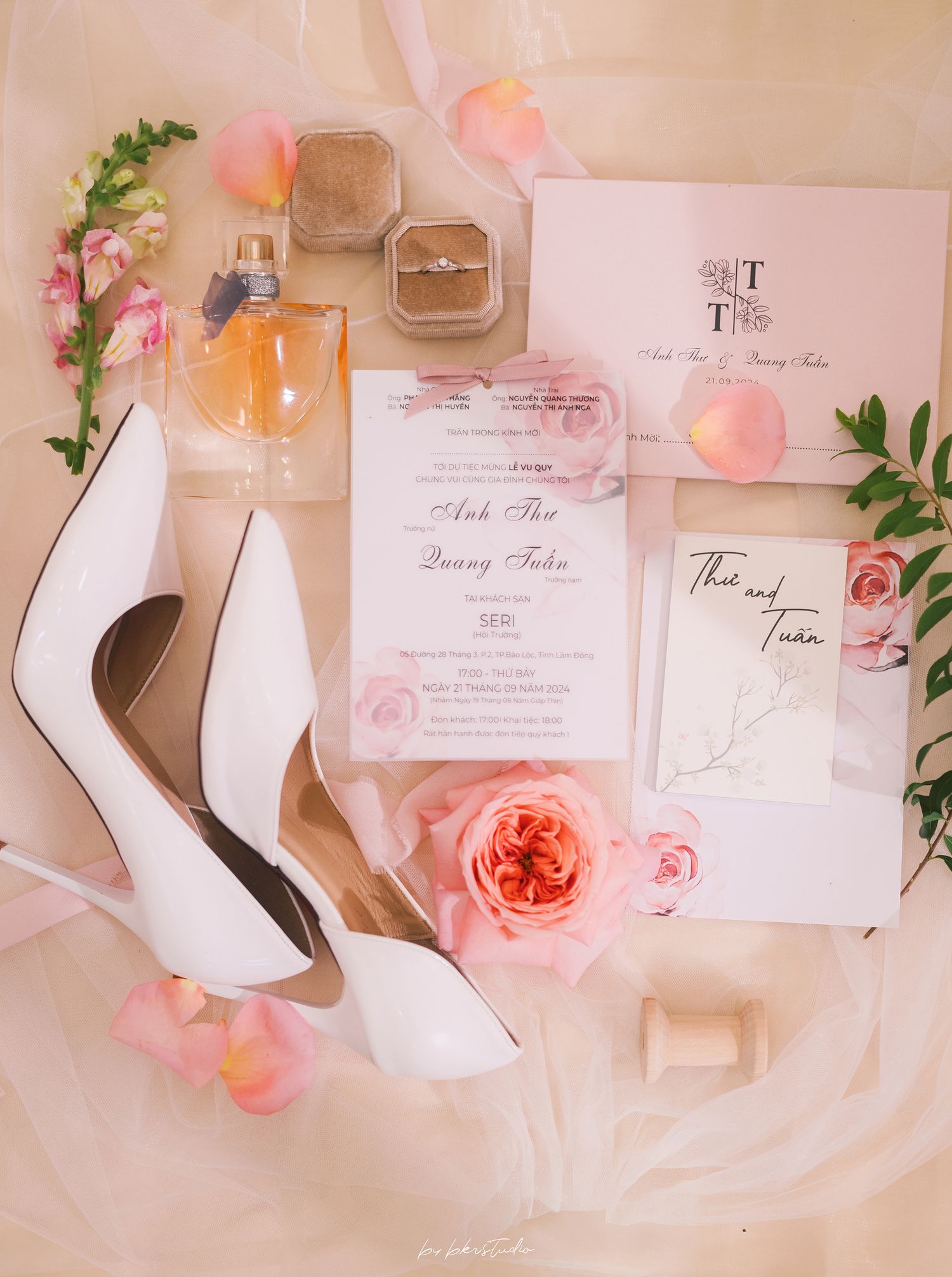 Wedding details flat lay with white heels, pink roses, invitations and perfume bottles on light fabric background.