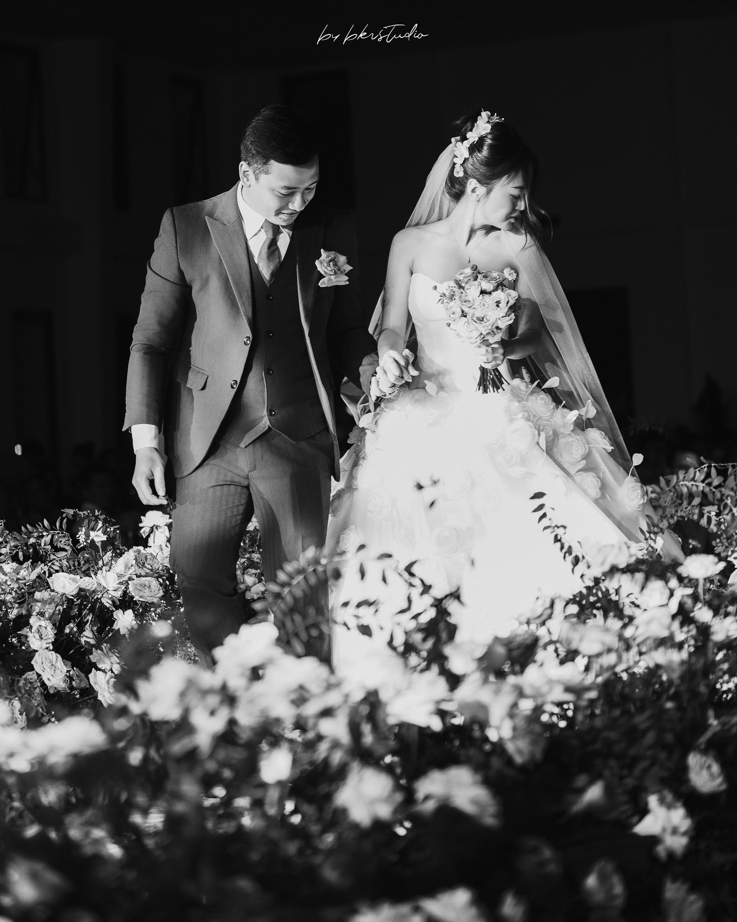 Black and white wedding photo of a couple seated among flowers, with the bride wearing a veil and traditional white gown.