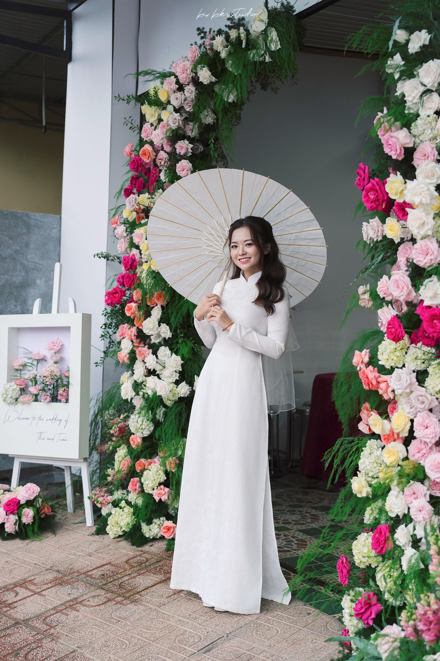 A traditional white ao dai dress is worn by someone posing with a parasol against a pink and white floral arch backdrop.