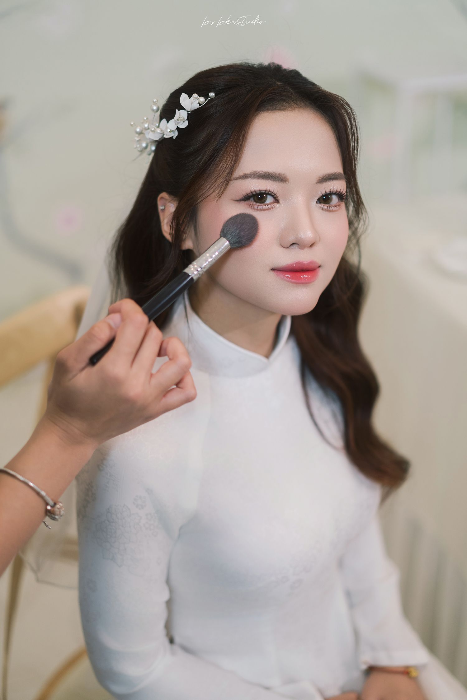 A person in a white turtleneck sweater getting makeup applied with a brush while wearing delicate hair accessories.