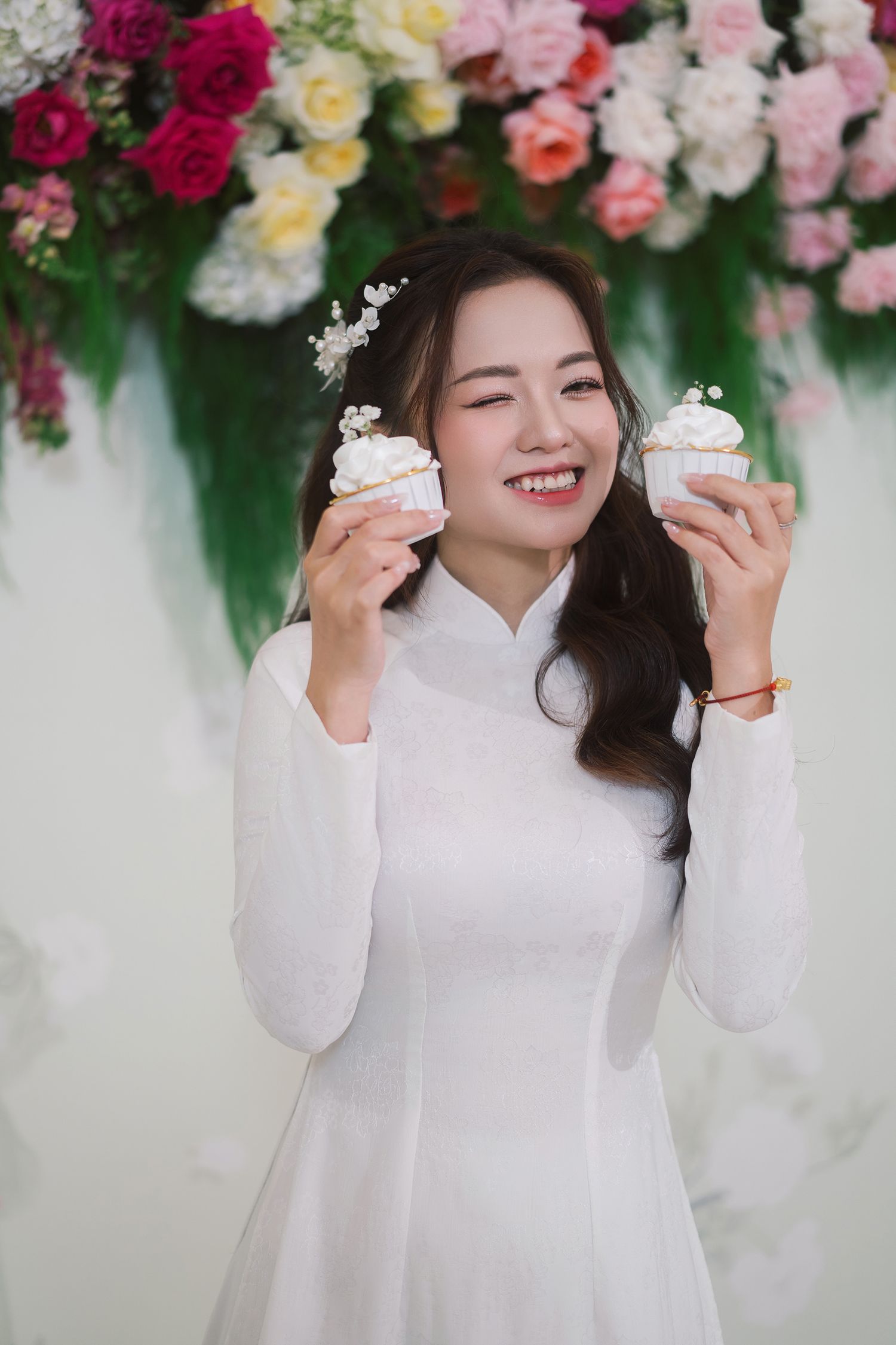 A person wearing a traditional white ao dai dress holds two cupcakes while smiling in front of a floral backdrop.