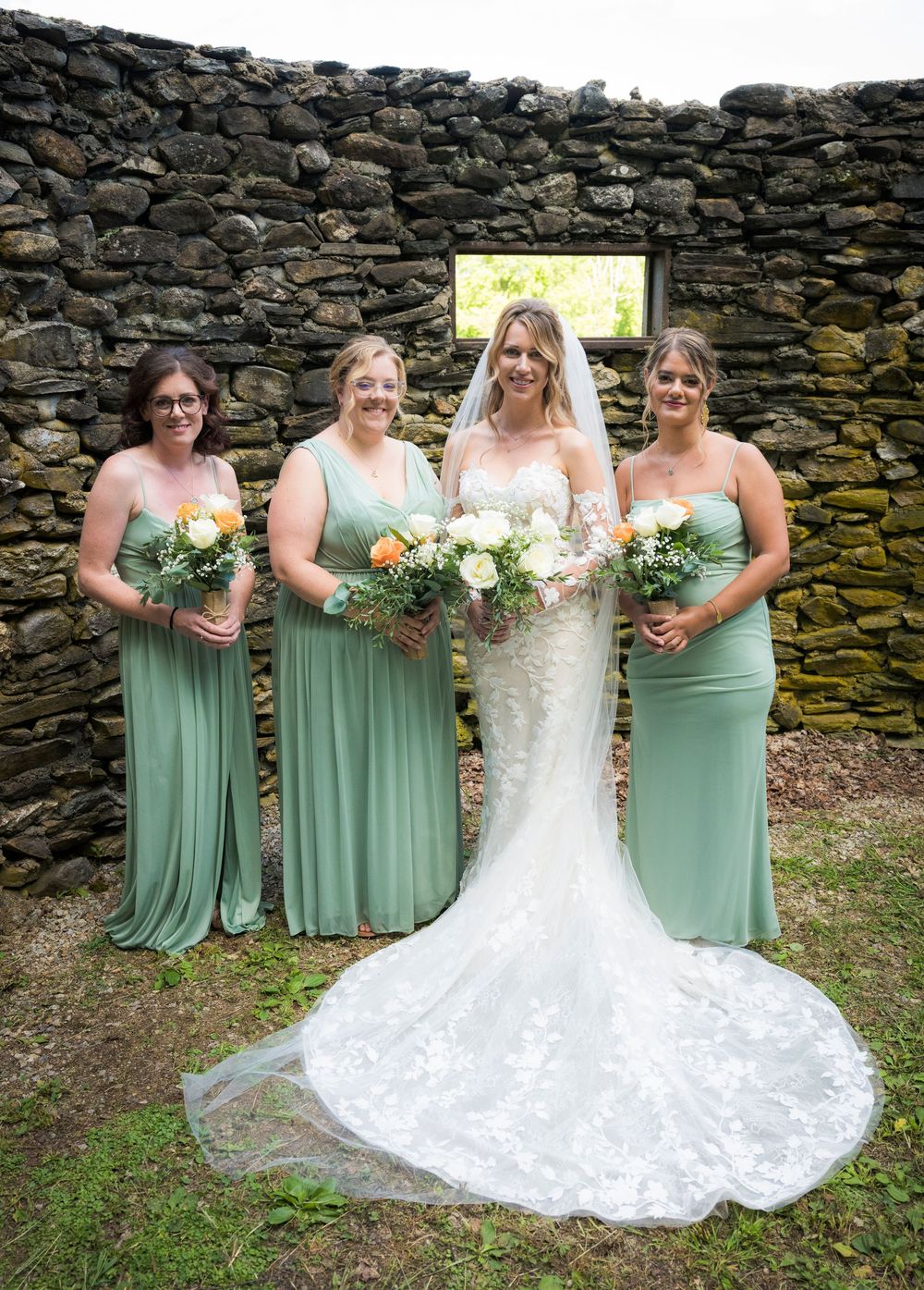 Wedding party poses against rustic stone wall with bridesmaids in sage green dresses and bride in lace gown.