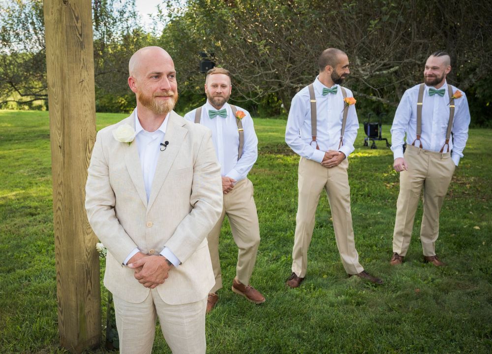 A panoramic view of a wedding party in light beige suits and suspenders standing casually in a green outdoor setting.